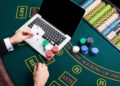 A person is holding two aces, a club, and a spade, in front of a laptop on a poker table covered with various poker chips and cards. The table is marked with areas labeled "BET" and "ANTE." The green felt table suggests a casino game setting. | MyBlackChip.com