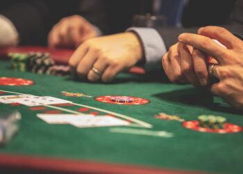 Close-up of people’s hands at a casino table playing cards, with poker chips and cards visible on a green felt surface. The players are wearing suits, and the focus is on the game in progress. | MyBlackChip.com