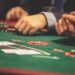 Close-up of people’s hands at a casino table playing cards, with poker chips and cards visible on a green felt surface. The players are wearing suits, and the focus is on the game in progress. | MyBlackChip.com