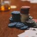 A close-up of assorted poker chips stacked on a wooden table next to playing cards and a blurred glass of brown liquid in the background. | MyBlackChip.com