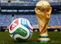 A colorful FIFA-approved soccer ball and the gold FIFA World Cup trophy are placed on a green field inside a large, empty stadium. | MyBlackChip.com